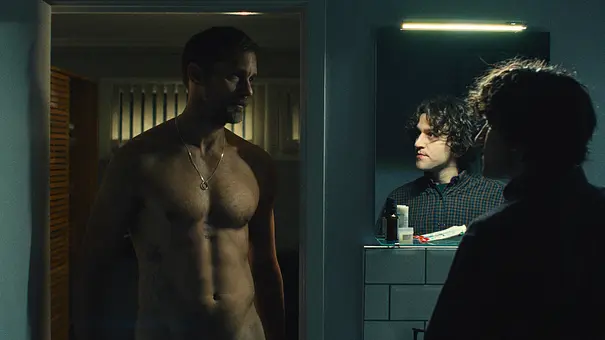 Alexander Skarsgård steht nackt vor Harry Melling, der vor einem Spiegel steht und Skarsgård überwältigt ansieht. - Foto: Element Pictures PLN Limited, British Broadcasting Corporation and the British Film Institute Alexander Skarsgård steht nackt vor Harry Melling, der vor einem Spiegel steht und Skarsgård überwältigt ansieht. - Foto: Element Pictures PLN Limited, British Broadcasting Corporation and the British Film Institute