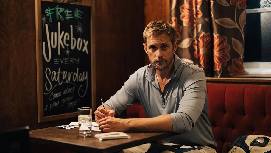 Alexander Skarsgård sitzt an einem Tisch in einer Bar und macht sich Notizen. Neben ihm ist ein Schild, auf dem unter anderem Jukebox steht. 
