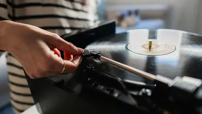 Ein Plattenspieler in der Nahansicht, während der Tonarm von einer weiblichen Hand auf dem Abspielmedium platziert wird. - Foto: iStock/ Ivan Rodriguez Alba
