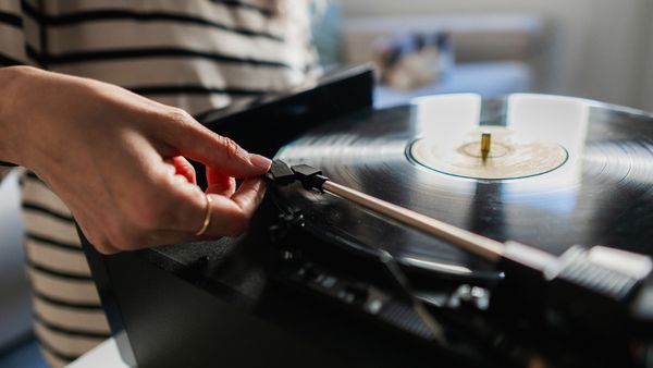Ein Plattenspieler in der Nahansicht, während der Tonarm von einer weiblichen Hand auf dem Abspielmedium platziert wird. - Foto: iStock/ Ivan Rodriguez Alba