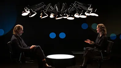 Zwei Personen sitzen sich in einem dunklen Studio gegenüber und führen ein Gespräch. Zwischen ihnen steht ein runder, beleuchteter Tisch, über ihnen hängt eine Gruppe von Studiolampen. Der Hintergrund ist schwarz mit dezenten blauen Lichtpunkten. - Foto: ZDF / Juliane Erich