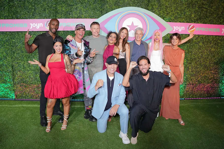 Die Promi Big Brother Kandidaten 2026 posieren vor einer grünen Wand mit „Promi Big Brother“-Logo. Zwei Männer knien vor der Gruppe.
