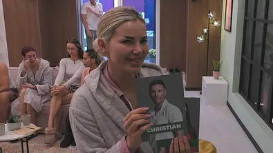 Kim Virginia Grey hält bei PromiBB im grauen Bademantel eine Porträtkarte mit der Aufschrift „Christian in die Kamera, um ihn rauszuwählen. - Foto: Joyn Kim Virginia Grey hält bei PromiBB im grauen Bademantel eine Porträtkarte mit der Aufschrift „Christian in die Kamera, um ihn rauszuwählen. - Foto: Joyn