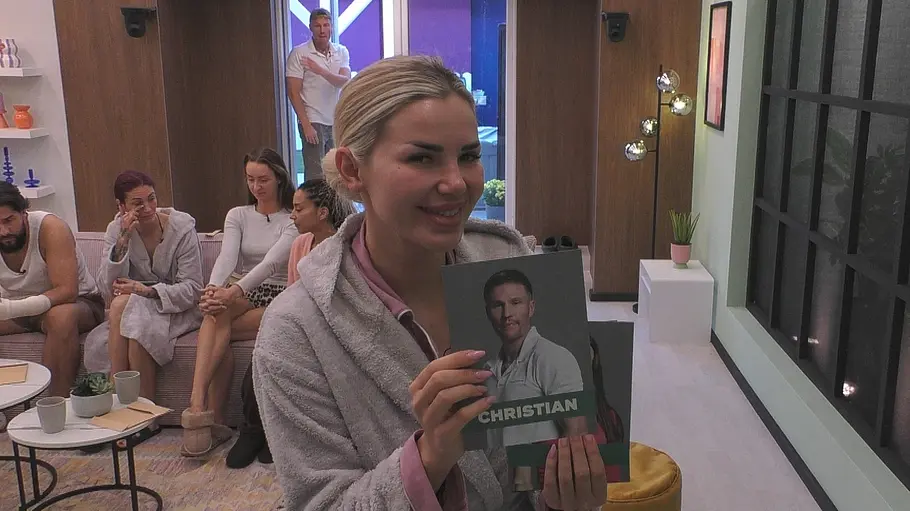 „Promi Big Brother 2026, Tag 9: Wer ist raus? Kim Virginia Grey hält bei PromiBB im grauen Bademantel eine Porträtkarte mit der Aufschrift „Christian in die Kamera, um ihn rauszuwählen.
