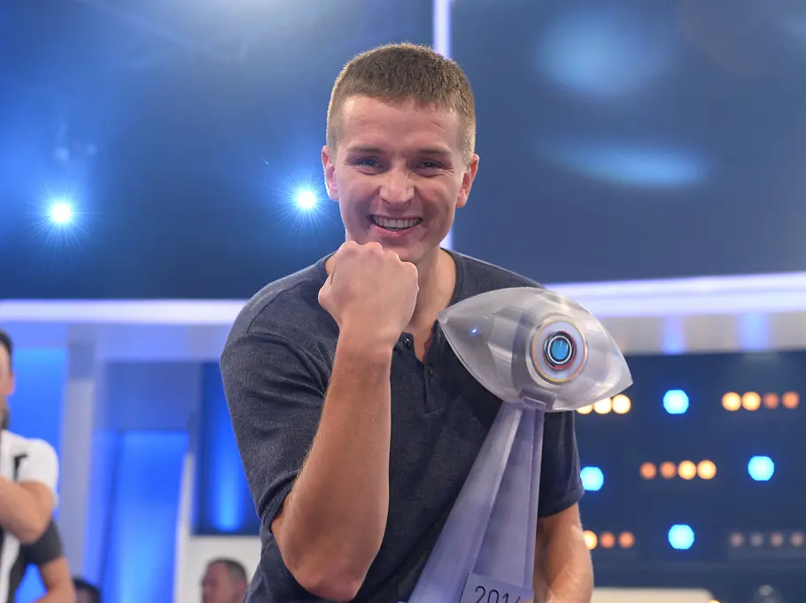 Promi Big Brother-Gewinner 2014 – Aaron Troschke Aaron Troschke lächelt mit erhobener Faust und einer augenförmigen Trophäe auf einer blau beleuchteten Bühne.