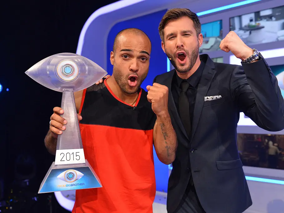 Promi Big Brother-Gewinner 2015 – David Odonkor David Odonkor hält einen augenförmigen Pokal mit der Jahreszahl 2015 und jubelt mit dem PromiBB-Moderator im dunklen Anzug mit erhobener Faust.