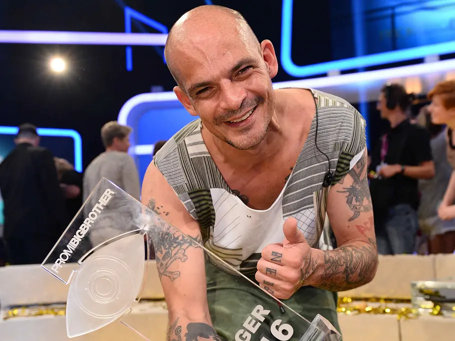 Promi Big Brother-Gewinner 2016 – Benjamin Tewaag Benjamin Tewaag hält lächelnd den „Promi Big Brother“-Siegerpokal 2016 und zeigt den Daumen hoch. Am Boden liegt Goldkonfetti.