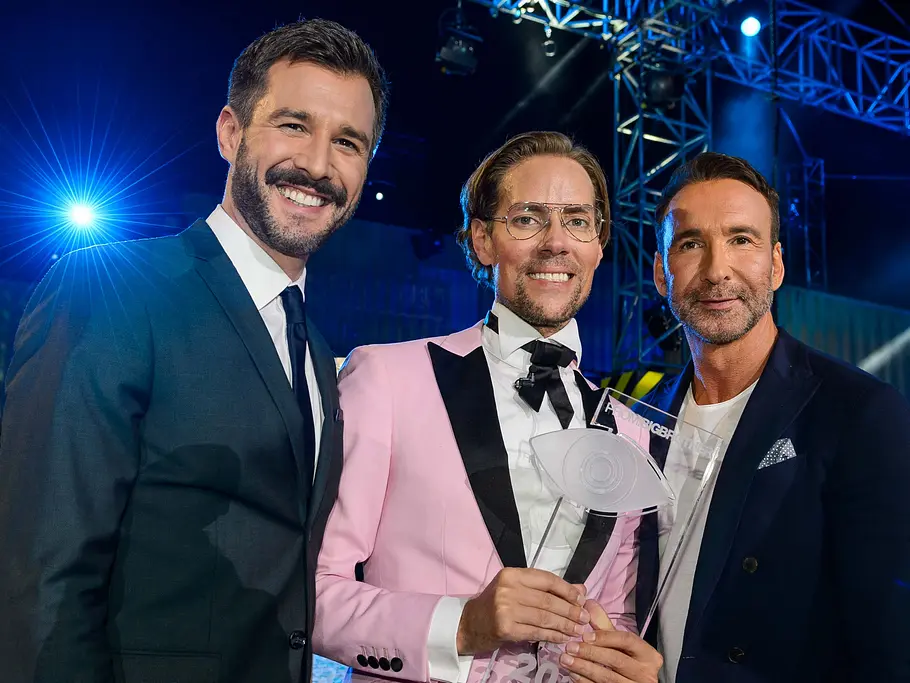 Promi Big Brother-Gewinner 2017 – Jens Hilbert Jens Hilbert steht im rosa Anzug mit einer transparenten Trophäe zwischen den Promi-BB-Moderatoren auf einer blau beleuchteten Bühne.