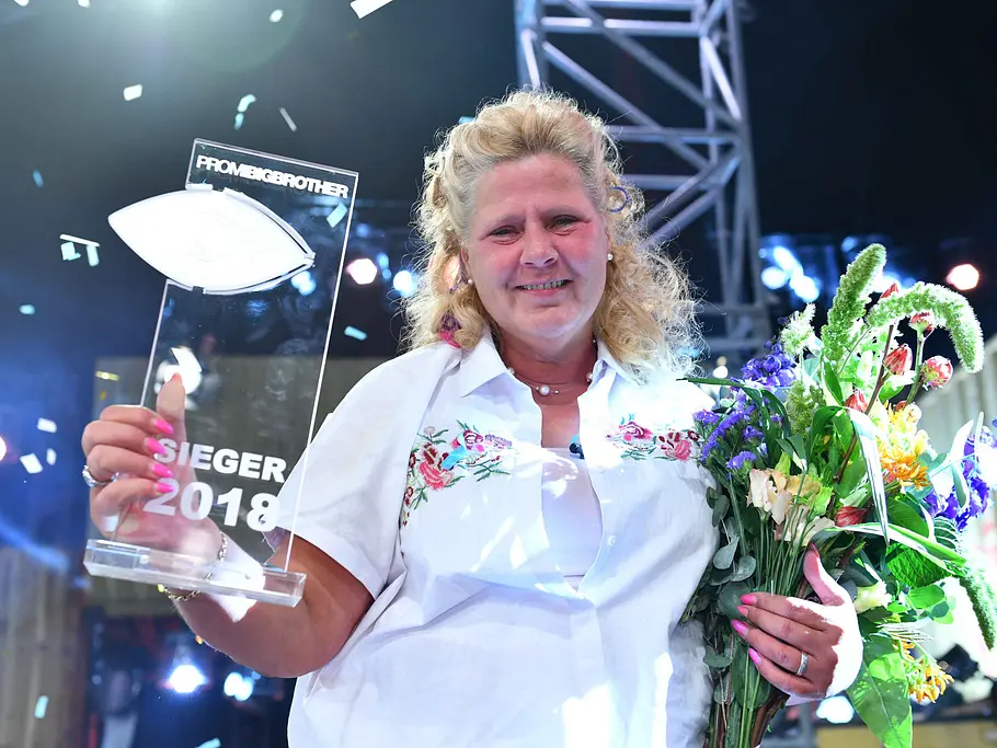Promi Big Brother-Gewinner 2018 – Silvia Wollny Silvia Wollny hält lächelnd den „Promi Big Brother“-Siegerpokal 2018 und einen Blumenstrauß, während Konfetti fällt.