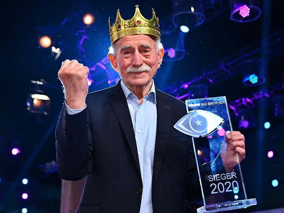 Promi Big Brother-Gewinner 2020 – Werner Hansch Werner Hansch trägt eine goldene Krone, hebt die Faust und hält den Siegerpokal von „Promi Big Brother“ 2020.