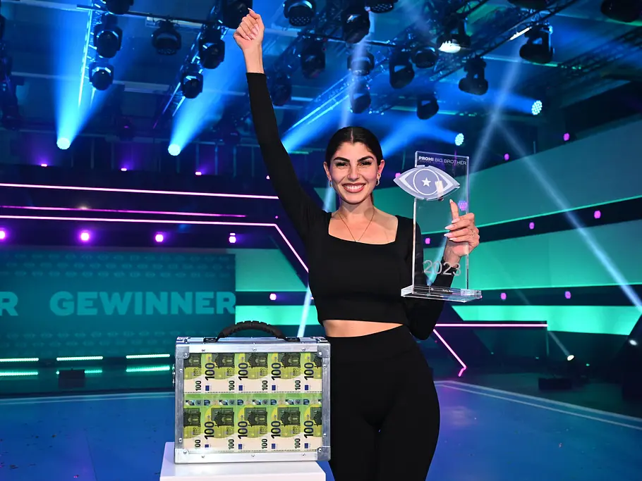 Promi Big Brother-Gewinnerin 2023 – Yeliz Koc Yeliz Koc hält auf einer bunt beleuchteten Bühne einen Pokal und hebt einen Arm. Neben ihr steht ein Koffer mit Geldscheinen.