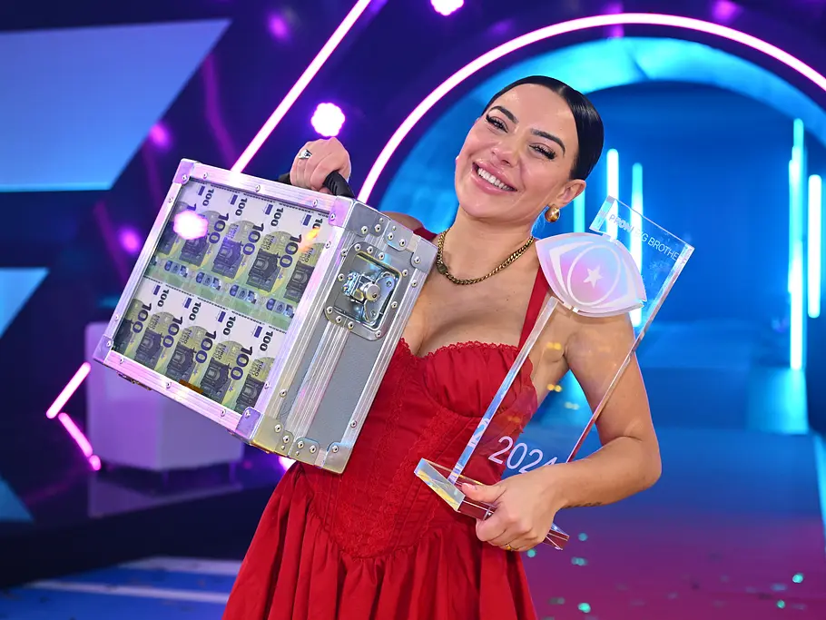 Promi Big Brother-Gewinner 2024 – Leyla Lahouar Leyla Lahouar lächelt im roten Kleid und hält einen Koffer voller Geldscheine sowie eine transparente Trophäe auf einer Bühne.