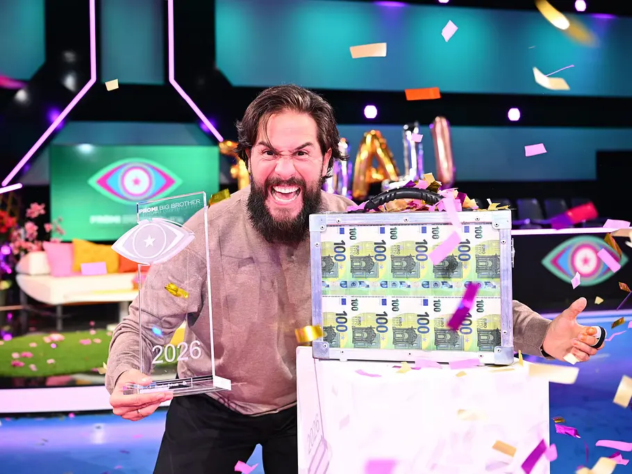 Promi Big Brother-Gewinner 2026 – Maurice Dziwak PromiBB-Gewinner Maurice Dziwak jubelt im Konfettiregen mit einer transparenten Trophäe neben einem Koffer voller 100-Euro-Scheine.