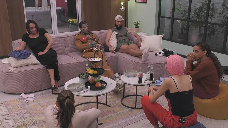 Die Promi Big Brother-Kandidaten 2026 sitzen in Folge 5 im Wohnzimmer um zwei Couchtische mit Obst und Getränken und reden.