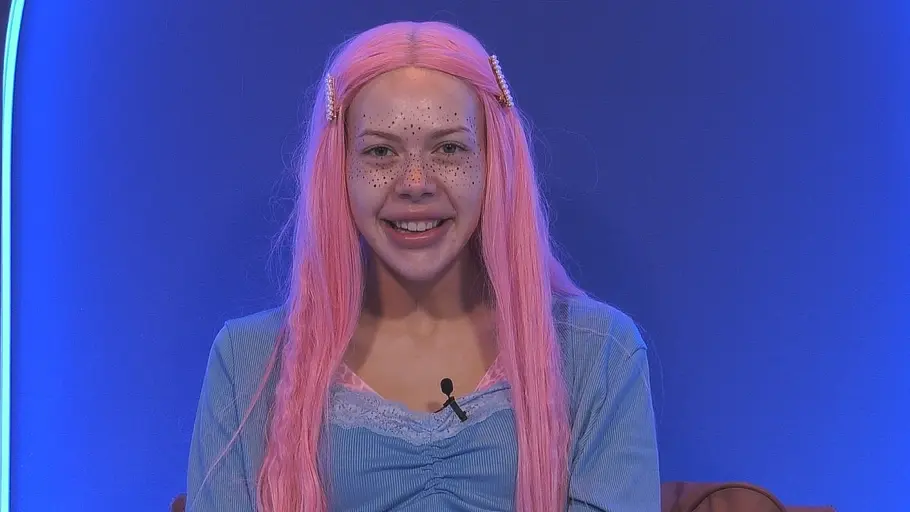 Nina Bridney Nina Bridney lächelt mit langen rosa Haaren, Haarspangen und dunklen Punkten im Gesicht vor blau beleuchtetem Hintergrund.