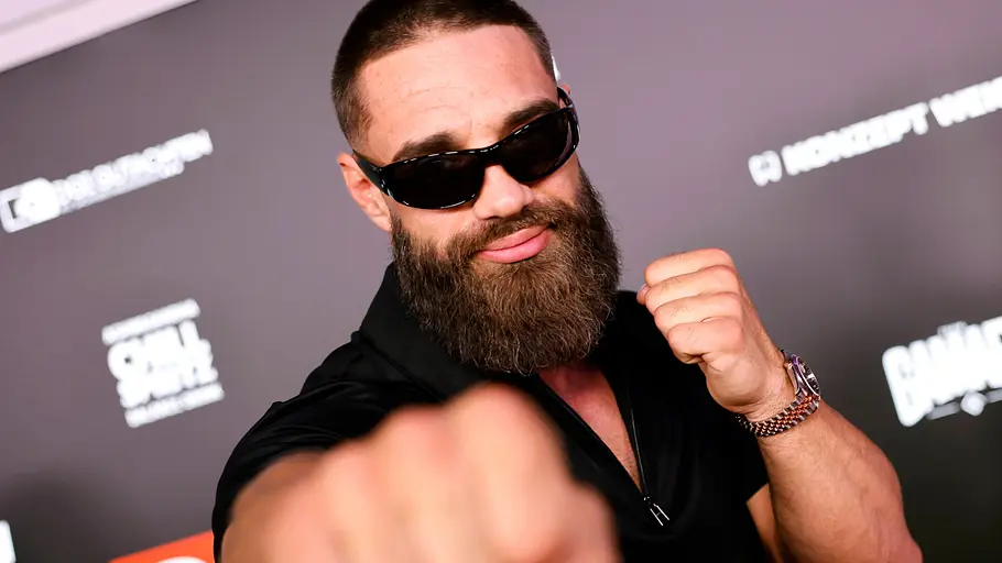 Aleks Petrovic Ein Mann mit dichtem Bart und kurzer Frisur posiert vor einer schwarzen Event-Wand mit verschiedenen Logos. Er trägt eine schwarze Sonnenbrille und ein schwarzes Oberteil. Mit einer Faust streckt er sich kämpferisch in Richtung Kamera, während die andere Hand ebenfalls zur Faust geballt ist. Durch die Perspektive wirkt die ausgestreckte Faust besonders groß im Vordergrund.