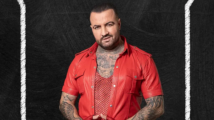Cosimo Porträt eines Mannes mit kurzen dunklen Haaren und Bart, der vor einem schwarzen Hintergrund in Kreidetafel-Optik posiert. Er trägt ein komplett rotes Outfit mit kurzer Jacke, Netzshirt und Handschuhen. Auffällige Tattoos bedecken seinen Hals und seine Arme.