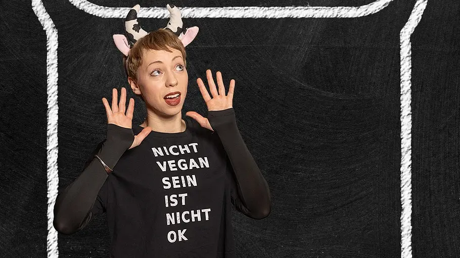 Raffaela Raab Porträt einer jungen Frau vor einem schwarzen Hintergrund in Kreidetafel-Optik. Sie trägt ein schwarzes Oberteil mit der Aufschrift „Nicht vegan sein ist nicht OK“, bordeauxrote Leggings und ein Haarreif mit Kuhhörnern und Ohren. Mit überraschter Mimik und erhobenen Händen posiert sie verspielt vor einer großen, mit weißer Kreide gezeichneten Sprechblase