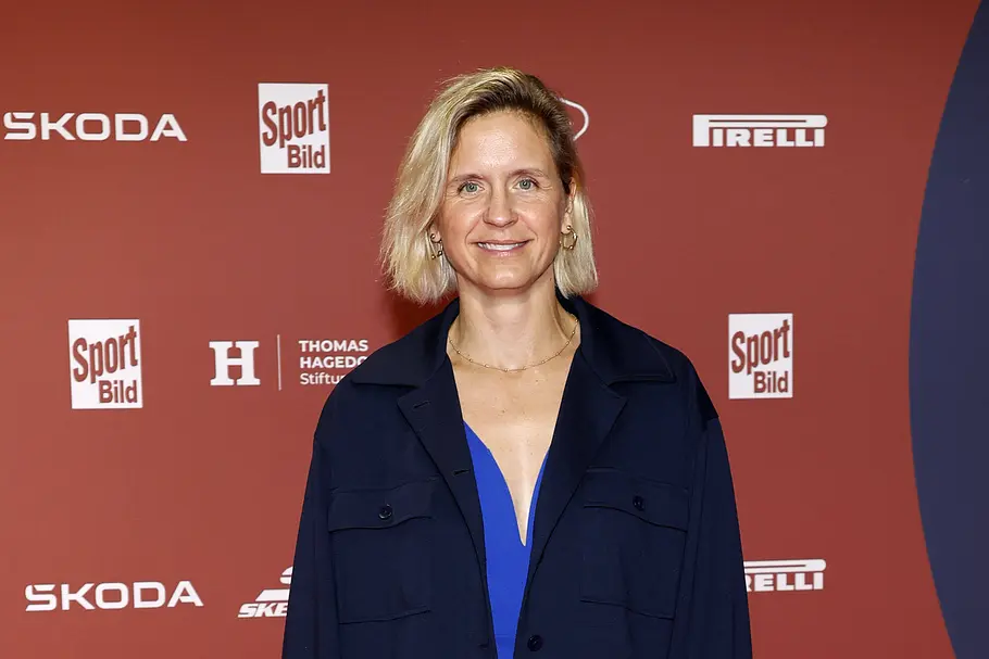 „Promi Padel WM“ 2026: Laura Ludwig Laura Ludwig steht lächelnd in einer dunkelblauen Jacke über einem blauen Oberteil vor einer roten Wand mit Sponsorenlogos.