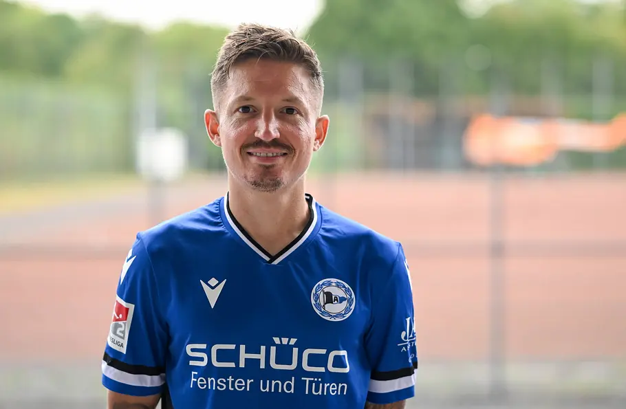 „Promi Padel WM“ 2026: Marc Rzatkowski Marc Rzatkowski lächelt im blauen Trikot von Arminia Bielefeld in die Kamera. Im Hintergrund ist ein Sportplatz zu sehen.