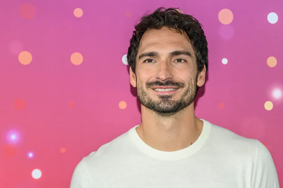 „Promi Padel WM“ 2026: Mats Hummels Mats Hummels lächelt in einem weißen Rundhalspullover vor einem pinkfarbenen Hintergrund mit bunten Punkten.