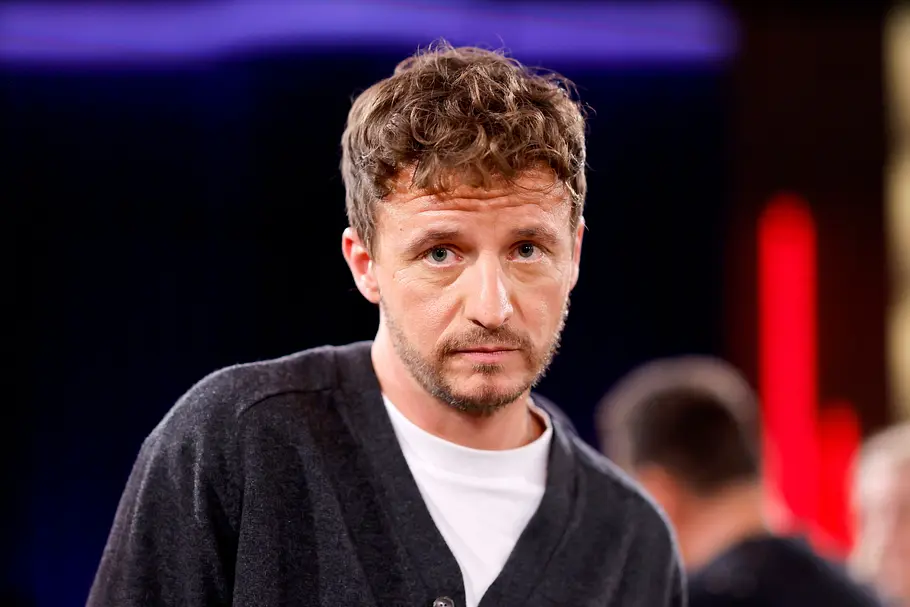 „Promi Padel WM“ 2026: Tommi Schmitt Tommi Schmitt trägt eine dunkelgraue Strickjacke über einem weißen T-Shirt vor einem unscharfen, farbig beleuchteten Hintergrund.