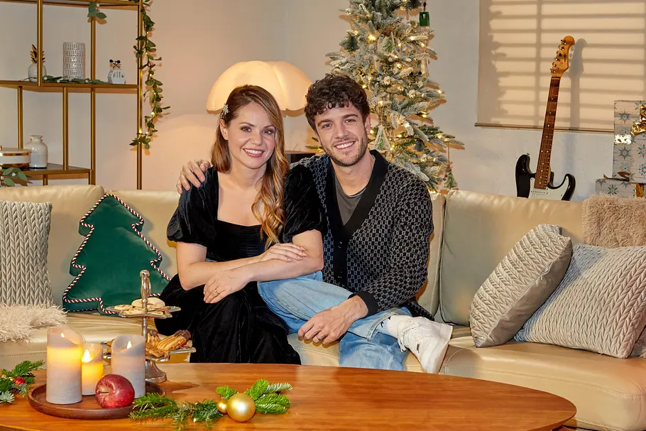Ein lächelndes Paar sitzt eng beieinander auf einem Sofa in einem gemütlich dekorierten Wohnzimmer. Im Hintergrund stehen ein geschmückter Weihnachtsbaum und warme Lichter, auf dem Tisch liegen Kerzen, Gebäck und weihnachtliche Dekoration – eine stimmungsvolle, festliche Szene.