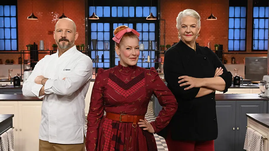 Promibacken Enie van de Meiklokjes steht zwischen den Juroren Christian Hümbs und Bettina Schliephake-Burchardt im Studio von „Das große Promibacken“ und alle blicken selbstbewusst in die Kamera.