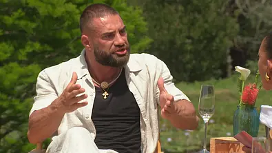Aleks  sitzt im Freien an einem Tisch und gestikuliert mit erhobenen Händen während eines offenbar emotionalen Gesprächs; vor ihm stehen ein leeres Sektglas und eine Vase mit Blumen. - Foto: RTL