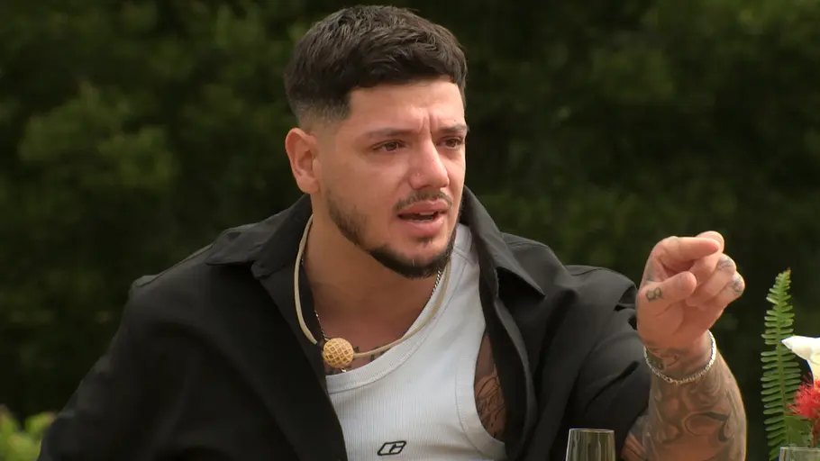 Gök Göker von Prominent getrennt Gök mit Tattoos zeigt mit ausgestrecktem Finger nach vorn und hat einen emotionalen Gesichtsausdruck. Er sitzt draußen vor grünem Hintergrund.