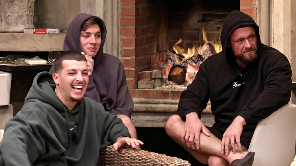Nico, Giuliano und Felix sitzen in der Villa vor dem Feuer, Nico lacht, die anderen beiden grinsen.