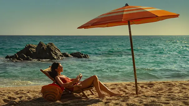 Frau im Badeanzug sitzt entspannt auf einem Liegestuhl unter einem orangefarbenen Sonnenschirm am Sandstrand, liest ein Buch und genießt den Blick aufs Meer, im Hintergrund Felsen im Wasser. - Foto: Daniel Escale/Netflix