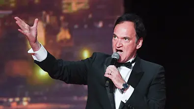 Quentin Tarantino steht im Anzug auf einer Bühne und spricht in ein Mikrofon. - Foto: IMAGO / ABACAPRESS Quentin Tarantino steht im Anzug auf einer Bühne und spricht in ein Mikrofon. - Foto: IMAGO / ABACAPRESS