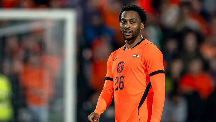 Fußballspieler der niederländischen Nationalmannschaft im orangefarbenen Trikot mit der Rückennummer 26 steht während eines Länderspiels auf dem Rasen und blickt lächelnd zur Seite. Im Hintergrund sind Tor und verschwommene Zuschauer zu sehen.