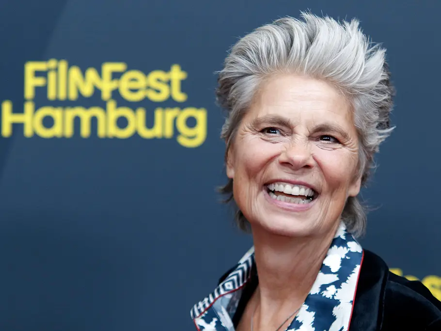 Quiz-Champion: Österreich-Deutschland-Special mit Sarah Wiener Sarah Wiener lächelt vor einer Wand mit dem Schriftzug „Filmfest Hamburg“. Sie trägt kurze graue Haare und einen gemusterten Kragen.