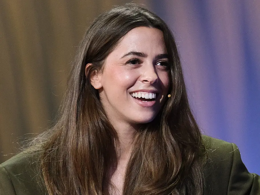 Quiz-Champion: Österreich-Deutschland-Special mit Vanessa Mai Vanessa Mai lächelt mit offenem Mund. Sie trägt einen dunkelgrünen Blazer und ein Headsetmikrofon zu langen braunen Haaren.