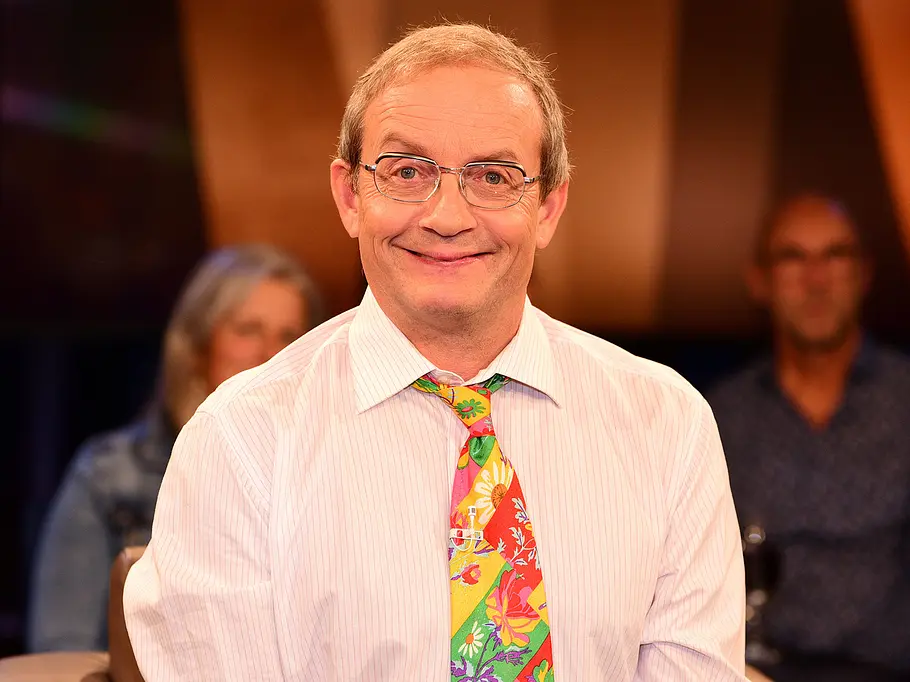 Quiz-Champion: Österreich-Deutschland-Special mit Wigald Boning Wigald Boning sitzt lächelnd mit Brille, hellem Hemd und bunter Blumenkrawatte vor unscharfem Studiopublikum.