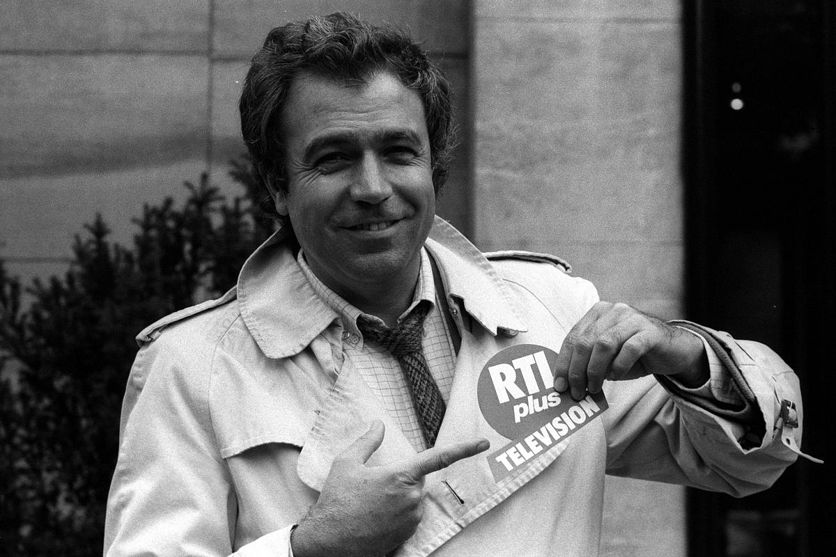 Rainer Holbe in den 1980er-Jahren, lächelnd im Trenchcoat, zeigt auf ein RTLplus-Logo – der Moderator war eines der prägenden Gesichter des deutschen Privatfernsehens.