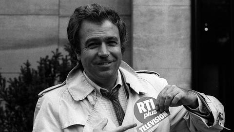 Rainer Holbe in den 1980er-Jahren, lächelnd im Trenchcoat, zeigt auf ein RTLplus-Logo – der Moderator war eines der prägenden Gesichter des deutschen Privatfernsehens. - Foto:  IMAGO / teutopress