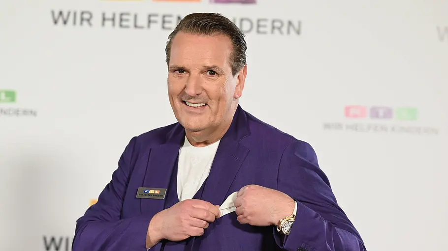 Ralf Dümmel Ralf Dümmel lächelt im violetten Anzug vor einer RTL-Logowand und richtet mit beiden Händen sein weißes Einstecktuch.