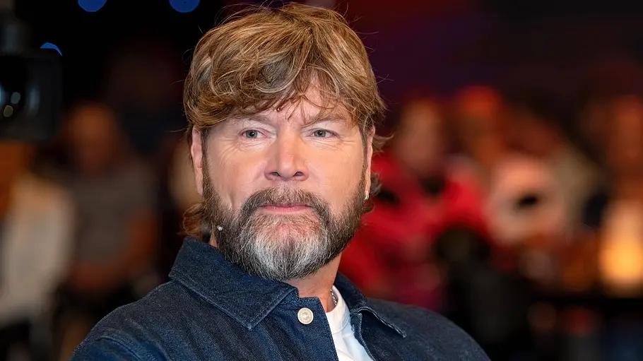 Rea Garvey trägt eine dunkelblaue Jacke über einem weißen Shirt. Im Hintergrund sind unscharf mehrere Menschen zu sehen.