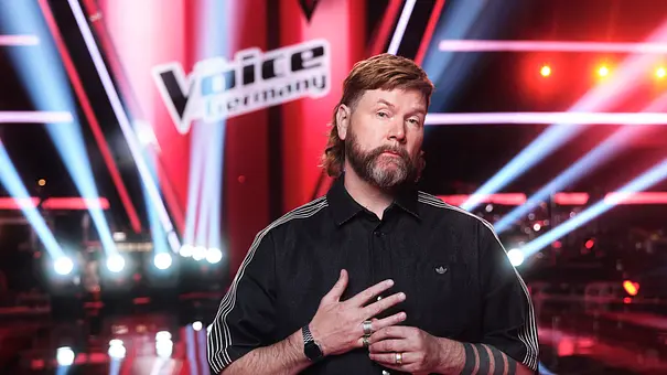 Coach Rea Garvey schaut monoton in die Kamera. Im Hintergrund sieht man das The Voice of Germany-Schild und viele Bühnenlichter. er trägt eine schwarze Adidas Jacke. - Foto: Seven.One Entertainment Group GmbH