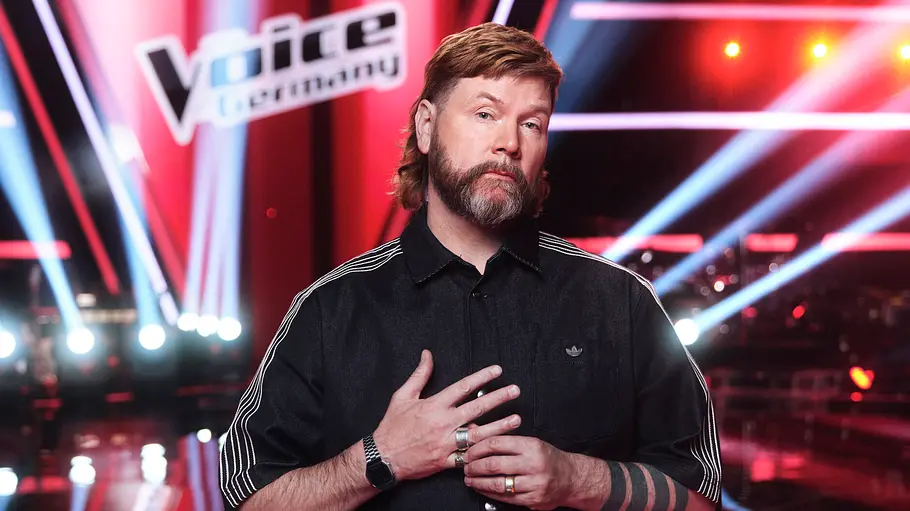 Rea Garvey The Voice of Germany Coach Rea Garvey schaut monoton in die Kamera. Im Hintergrund sieht man das The Voice of Germany-Schild und viele Bühnenlichter. er trägt eine schwarze Adidas Jacke.