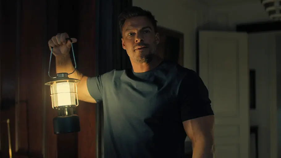 Reacher – Staffel 3 Alan Ritchson leuchtet mit einer Laterne ein dunkles Haus aus.