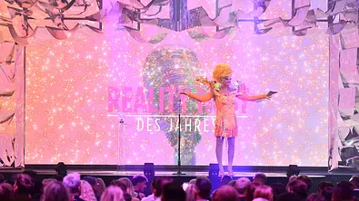 Olivia Jones in auffälligem Bühnenoutfit steht vor leuchtender Kulisse auf der Awards-Bühne. - Foto: RTL / Julia Feldhagen