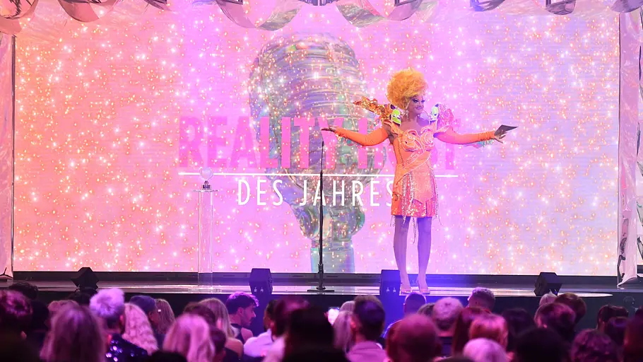 Olivia Jones bei den Reality Awards Olivia Jones in auffälligem Bühnenoutfit steht vor leuchtender Kulisse auf der Awards-Bühne.