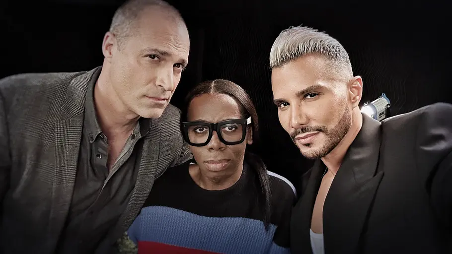 Juroren von ANTM Drei Personen posieren eng nebeneinander vor dunklem Hintergrund und blicken direkt in die Kamera. Links steht ein Mann mit sehr kurzem, hellem Haar in einem grauen Sakko. In der Mitte eine Person mit großen, schwarzen Brillenfassungen und einem dunklen Pullover mit breiten farbigen Streifen. Rechts ein Mann mit kurz geschnittenem, hellblondem Haar und Bart, der ein schwarzes Sakko über einem hellen Oberteil trägt. Die Aufnahme wirkt wie ein Studio- oder Backstage‑Porträt mit gerichteter Beleuchtung.