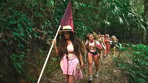 Eine Gruppe von Frauen wandert in einer Reihe durch einen dichten Dschungel. Die vorderste Frau trägt ein Sommerkleid und einen Strohhut und hält eine Fahne. Die anderen Frauen tragen sportliche Kleidung oder Outfits in Rosa und tragen Wanderrucksäcke. Der Pfad, auf dem sie laufen, ist schmal und von üppigem Grün umgeben. - Foto: RTL