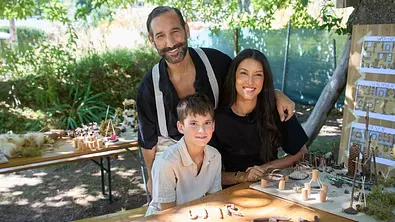 Rebecca Mir und Massimo Sinato stehen mit einem Jungen hinter einem Tisch mit Bastelarbeiten aus Naturmaterialien im Freien. - Foto: RTL / Bob van Ooik