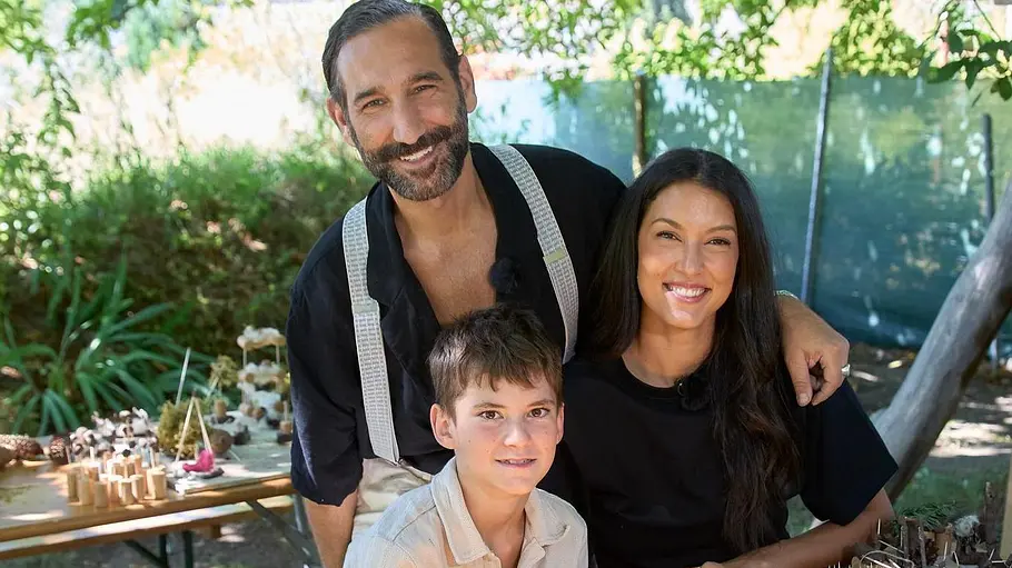 Rebecca Mir und Massimo Sinato Rebecca Mir und Massimo Sinato stehen mit einem Jungen hinter einem Tisch mit Bastelarbeiten aus Naturmaterialien im Freien.